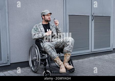 Behinderter Veteran im Rollstuhl, der Zigarette anzündet und eine Flasche Alkohol im Freien hält Stockfoto