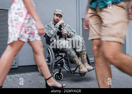 Betrunkener behinderter Soldat, der im Rollstuhl sitzt, mit Flasche und Zigarette in der Nähe verschwommener Fremder, die vorbeikommen Stockfoto