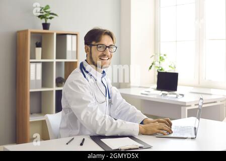 Porträt eines glücklichen jungen männlichen Arztes, der am Laptop an seinem Arbeitsplatz im Krankenhausbüro sitzt. Stockfoto