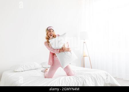 Lustige glückliche tausendjährige hübsche kaukasische blonde Frau in rosa Pyjama, Schlafmaske, umarmen Kissen, viel Spaß auf Komfort-Bett Stockfoto