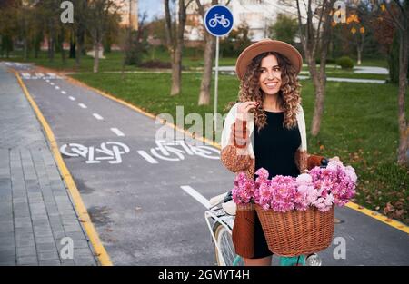 Lockige Frau, die auf einem Vintage-Fahrrad mit einem großen Korb mit rosa Blumen reitet. Lächelnde Frau mit braunem Hut und Strickjacke auf dem Fahrrad mit chrysanths am Lenker. Konzept eines gesunden Lebensstils. Stockfoto