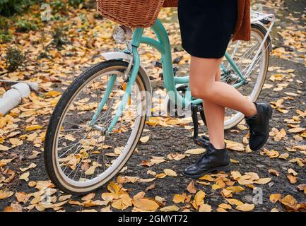 Nahaufnahme von weiblichen Beinen in eleganten Schuhen und blauem Fahrrad auf der Herbststraße. Junge Frau, die mit dem Fahrrad auf Asphalt mit gelben Blättern steht und Zeit im Freien verbringt. Stockfoto