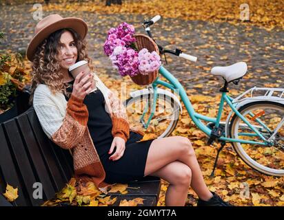 Seitenansicht einer jungen Frau mit lockigen Haaren im Hut, die auf einer holzbraunen Bank in der Nähe des Fahrrads sitzt und im Stadtpark leckeren heißen Kaffee oder Tee trinkt. Konzept der Ruhepause der frischen Luft im Herbst. Stockfoto
