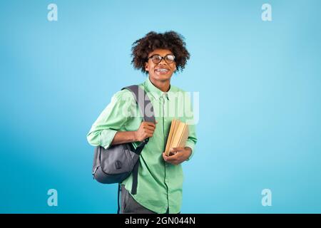 Fröhlicher schwarzer Student mit Rucksack und Büchern, die über dem blauen Studiohintergrund die Kamera anlächeln Stockfoto