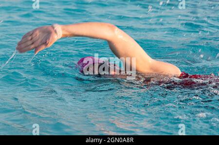 Nahaufnahme einer Frau, die in einem Pool schwimmt, wobei ihr Arm in der Luft hochgezogen ist, um im Freistil zu schwimmen, wobei sie eine pinke Mütze und Stacheln trägt. Stockfoto