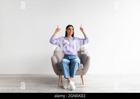 Lächelnde Frau, die im Sessel sitzt, mit beiden Händen nach oben zeigt und über ihrem Kopf leeren Raum bietet, Nachbau Stockfoto