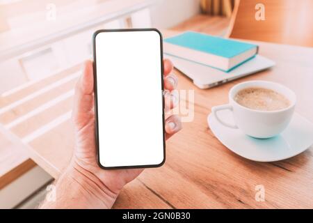 Leerer Smartphone-Bildschirm Mann hält Smartphone mit Kaffeetasse, Laptop-Computer und einem Buch im Heimbüro über den Schreibtisch, selektiver Fokus Stockfoto