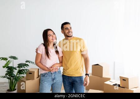 Glückliches junges multirassisches Paar, das am Umzugstag in ihrem Haus zwischen Kartons steht und Platz kopiert Stockfoto