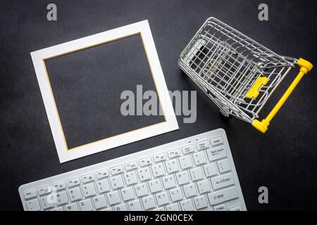 Weiße Computertastatur und Rahmen für Ihren eigenen Text, schwarzer Hintergrund, Miniatur eines Warenkorbs, Business- und Geschäftskonzept Stockfoto