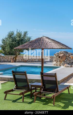 Zwei Sonnenliegen oder Sonnenliegen unter einem Regenschirm im tropischen Stil aus Bast oder Korbweide an der Seite eines Swimmingpools in einer privaten Ferienvilla auf zakynthos. Stockfoto