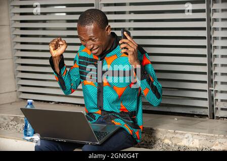 Ein hübscher junger afrikanischer Mann, der glücklich und aufgeregt mit einem Laptop sitzt und arbeitet, mit erhobenen Armen eine Geste macht, lächelt und nach Erfolg schreit Stockfoto