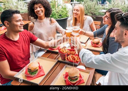 Eine Gruppe von multiethnischen, fröhlichen Freunden, die sich mit Fast Food und klirrenden Gläsern Bier am Tisch treffen, während sie Toast anbieten und den eines Freundes feiern Stockfoto