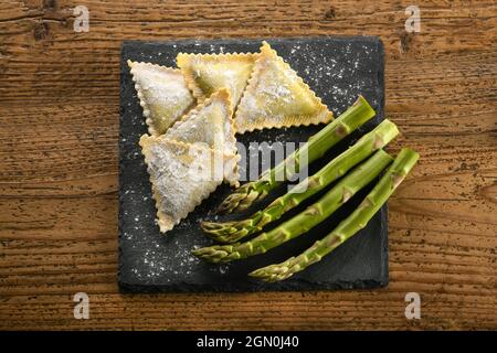 Von oben von rohen traditionellen italienischen dreieckigen Ravioli Pasta mit frischem grünen Spargel auf Schiefertafel bestreut mit Mehl auf Holz ta gelegt Stockfoto