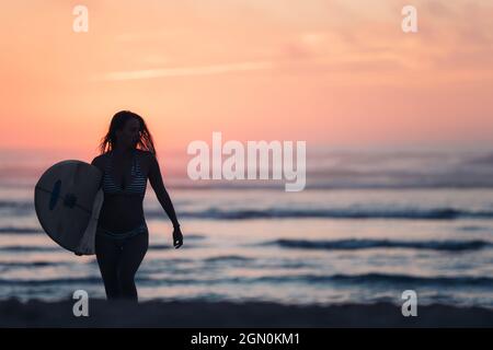 Weibliche Surferin geht mit Surfbrett am Strand in Sonnenuntergang, Surfen, Portugal, Sonnenuntergang Stockfoto