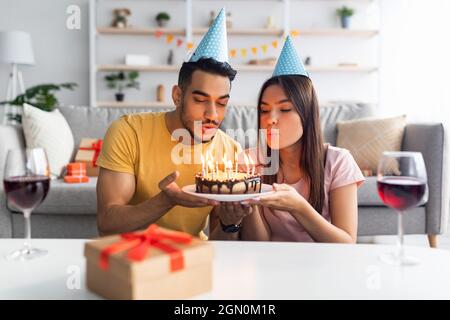 Positive interracial Paar in Party-Hüte blasen brennende Kerzen auf Geburtstagskuchen, Wein trinken, Geschenke zu Hause austauschen Stockfoto