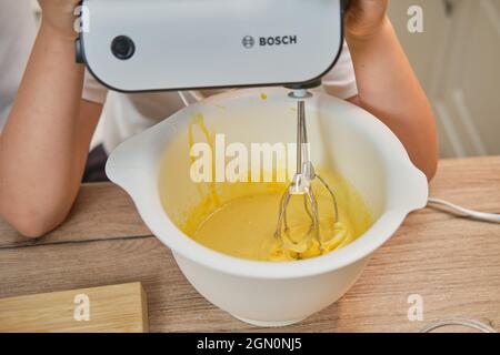 Junge in weißer Chefkleidung schlägt Eier mit einem Bosch-Mixer, um in seiner eigenen Küche einen Kuchen zu backen - Moskau, Russland, 2. September 2021 Stockfoto