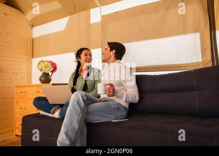 Fröhliches, multiethnisches Paar mit Tasse, das auf dem Laptop auf der Couch im Glamping House aufliegt Stockfoto