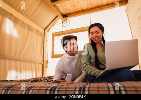 Multiethnisches Paar mit Gadgets auf dem Bett im Glamping-Haus Stockfoto