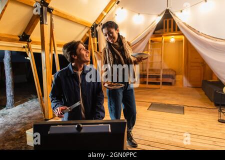 Mann, der auf der Terrasse des Glamping-Hauses die Freundin mit Essen auf den Tellern anschaut Stockfoto