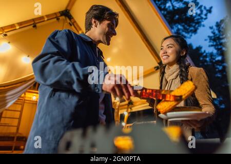 Low-Angle-Ansicht von lächelnden interracial paar Kochen auf Grill in der Nähe Glamping House am Abend Stockfoto