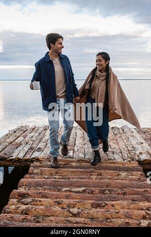 Lächelndes interrassisches Paar mit Decke und Tasse auf dem Pier in der Nähe des Meeres Stockfoto