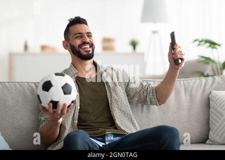 Positiver junger Araber, der Fußballspiele im Fernsehen ansieht, die Fernbedienung und den Ball zu Hause hält Stockfoto