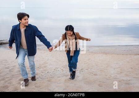 Lächelndes interrassisches Paar, das beim Gehen am Strand die Hände hält Stockfoto