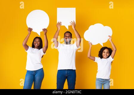 Glückliche schwarze Dreierfamilie in weißen T-Shirts, die leere Sprechblasen mit unterschiedlicher Form über ihren Köpfen halten und isoliert im gelben Studio stehen. Stockfoto