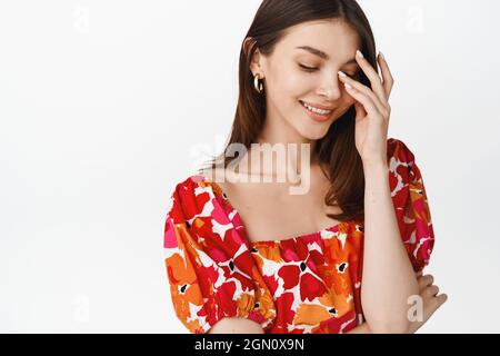 Schöne junge Frau lächelt kokettisch, berührt ihr Gesicht und schaut schüchtern erröten, stehen in Blumenkleid vor weißem Hintergrund Stockfoto