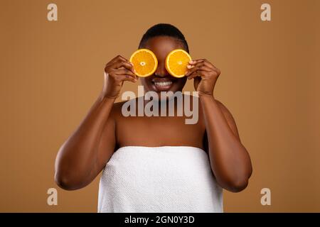 Hautpflegekonzept. Lustig lächelnde junge schwarze Frau, die Hälften von saftigen Zitrusfrüchten hält und die Augen bedeckt, Gläser aus Orange macht, isoliert über b Stockfoto