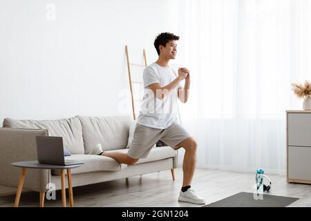 Online-Schulungen. Lächelnder Sportlicher Asiatischer Kerl, Der Einbeinige Geteilte Squats In Der Nähe Der Couch Macht, Indem Er Während Des Trainings Zu Hause Einen Laptop-Computer Verwendet. Inland M Stockfoto