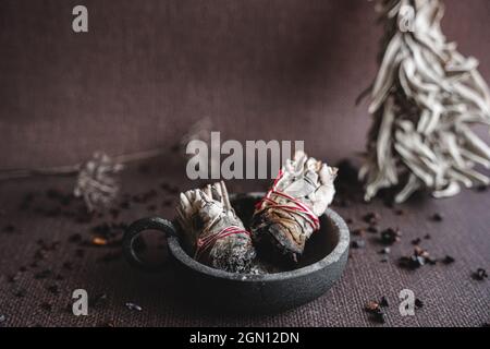 Zwei Salbei-Wischersticks in einer gusseisernen Schüssel. Dunkelbrauner Hintergrund mit weißem Salbeibündel und einigen getrockneten Kräutern im Hintergrund. Stockfoto