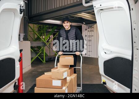 Lächelnder Kurier in Uniform, der im Lager steht und den Wagen für die Lieferung vorbereitet Stockfoto