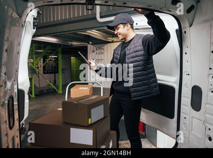Junger Mann, der ein digitales Tablet hält, während er am Van steht. Kurier überprüft Kartons auf dem Wagen. Stockfoto