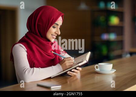 Schöne junge Frau im Hijab, die Notizen im Café schrieb Stockfoto