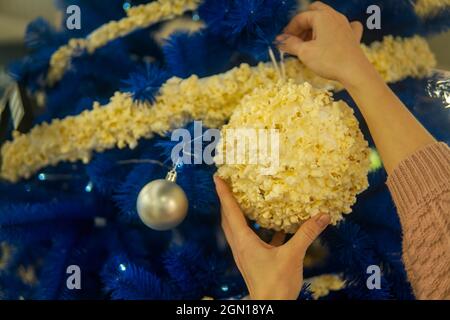Frauenhänden hängen einen großen Weihnachtsball mit Popcorn an einen blauen Weihnachtsbaum Stockfoto