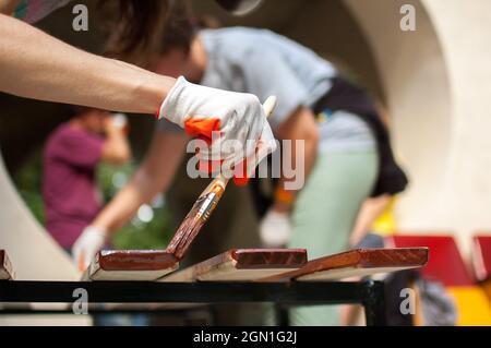 Die Arbeiterin macht die Malarbeiten aus den Holzprodukten, die Planke mit der braunen Farbe für die Herstellung der Bank, die Tischlerarbeiten, den Schutz des Holzes. Stockfoto