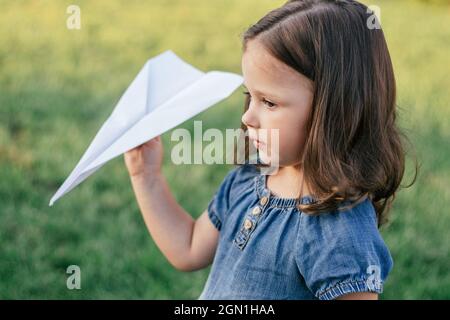 Kleines Mädchen 3-4 mit dunklen Haaren in Denim-Kleid in der Sonne startet Papierflugzeug auf grünem Rasen stehen Stockfoto