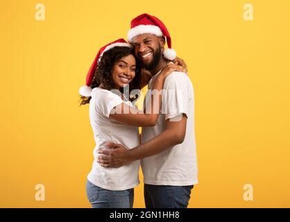 afroamerikanische Liebhaber in Weihnachtsmützen umarmen auf gelbem Studiohintergrund, feiern Neujahr 2022 oder Weihnachten Stockfoto