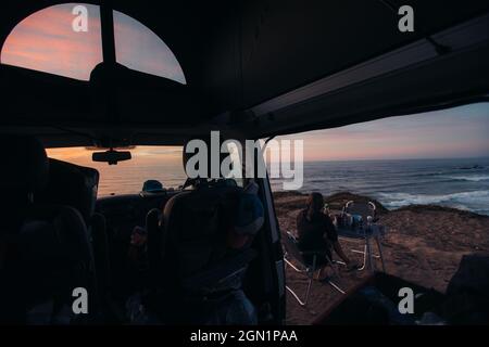 Frau sitzt vor ihrem Wohnmobil beim Frühstück mit der Küste von Portugal in Sicht, Urlaub, Camping, Portugal Stockfoto