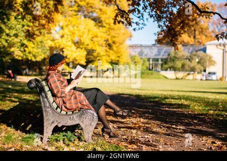 Rothaarige Frau im karierten Mantel und schwarzer Baskenmütze liest ein Buch auf der Bank und ruht sich an sonnigen Tagen im Herbstpark aus Stockfoto