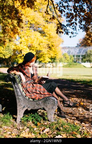 Rothaarige Frau in kariertem Mantel und schwarzem Baskenmütze, die auf der Bank liest und sich an sonnigen Tagen im Herbstpark ausruht Stockfoto