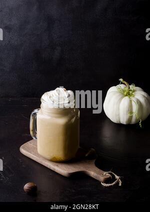 Würziger Kürbis-Latte in einem Glas mit Creme und Zimt. Gemütliches warmes Herbstgetränk-Konzept mit Kürbissen auf schwarzem Hintergrund in dunkler Tonart, selektiver Backgr Stockfoto