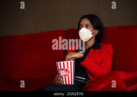 Junge afroamerikanische Frau im formellen Business-Anzug mit Schutzmaske im Kino. Sie isst ein Popcorn, während sie zuschaut - Covid-19 New Normal Stockfoto