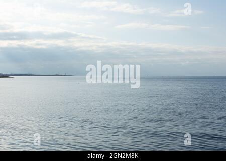 Stimmungsvolle Meereslandschaft. Blaue Wolken helle Strahlen der Sonne beleuchten die glatte Oberfläche des Meeres, des Ozeans. Panoramablick mit einem Leuchtturm Stockfoto