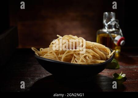 Appetitliche Spaghetti in Schale und grüne Basilikumblätter auf rustikalem Holztisch mit Olivenöl in Flasche in dunklem Studio serviert Stockfoto