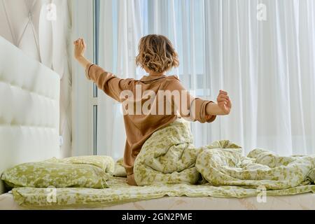 Rückansicht der jungen Frau, die die Arme auf dem sitzend streckt Bett zu Hause Schlafzimmer mit Blick auf das Fenster Stockfoto