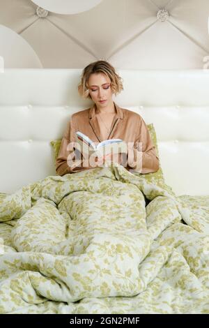 Clevere junge Frau sitzt im Bett unter der Decke mit Ein Buch in den Händen Stockfoto