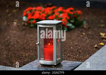 Grablampe aus Metall mit brennender Kerze auf einem Grab mit roten Blumen in verschwommenem Hintergrund Stockfoto