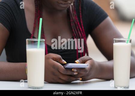 Zugeschnittenes Bild einer afroamerikanischen Frau, die ihr Telefon mit Milchshakes auf dem Tisch in einem Café im Freien benutzt Stockfoto
